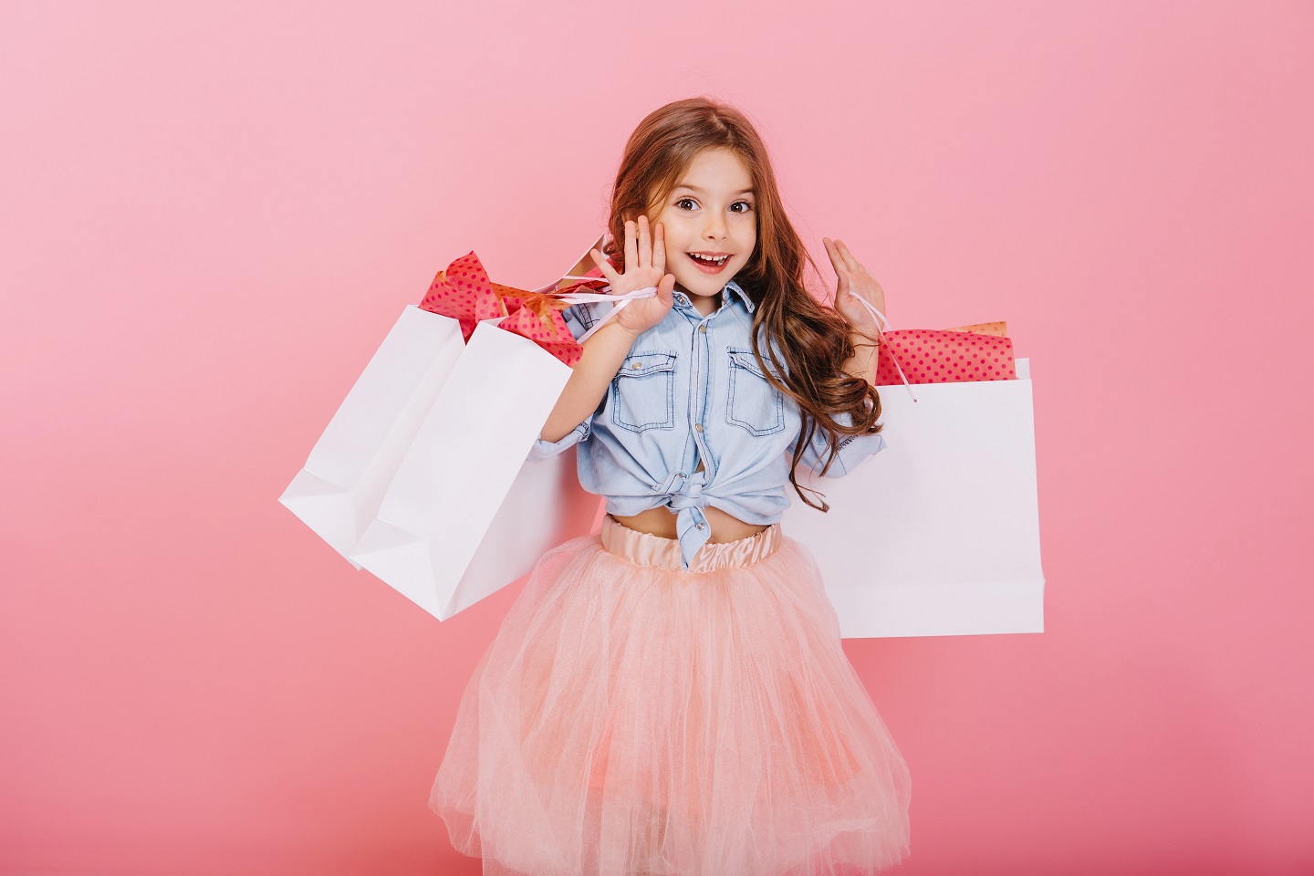Pretty joyful young girl in tulle skirt, with long brunette hair walking with white packages on pink background. Lovely sweet moments of little princess, pretty friendly child having fun to camera.